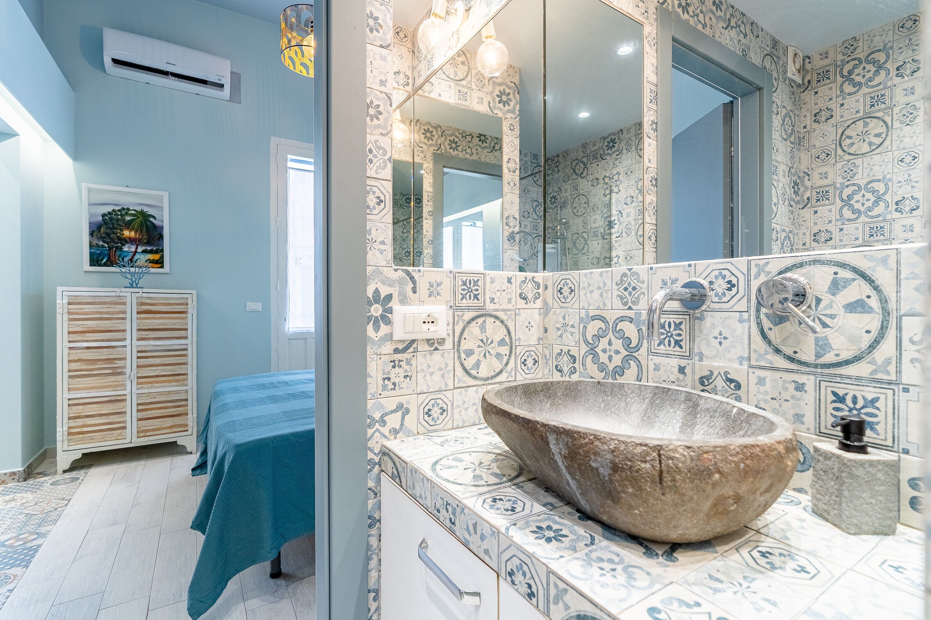 Une salle de bain avec un grand lavabo en pierre, des carreaux décorés, un miroir et un lit recouvert d’une courtepointe bleue.