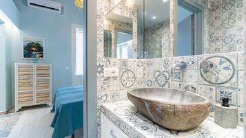 Une salle de bain avec un grand lavabo en pierre, des carreaux décorés, un miroir et un lit recouvert d’une courtepointe bleue.