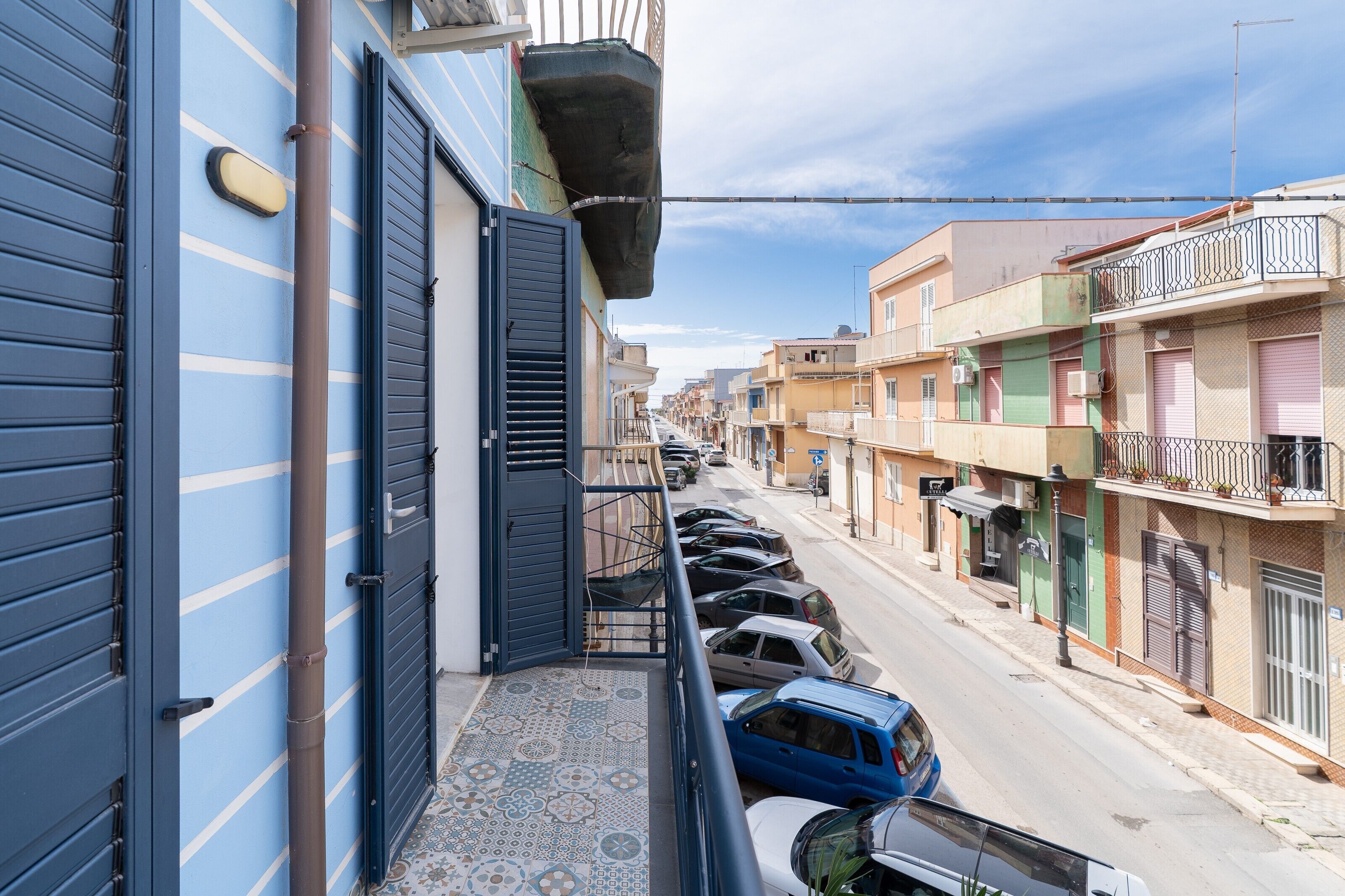 Eine Wohnstraße mit parkenden Autos, einem blauen Gebäude mit Fensterläden und einem Kachelbalkon.