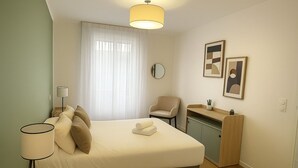Een keurig opgemaakt slaapkamer met een bed, nachtlampjes, een stoel, een klein bureau met een plant, en twee ingelijste kunstwerken aan de muur.