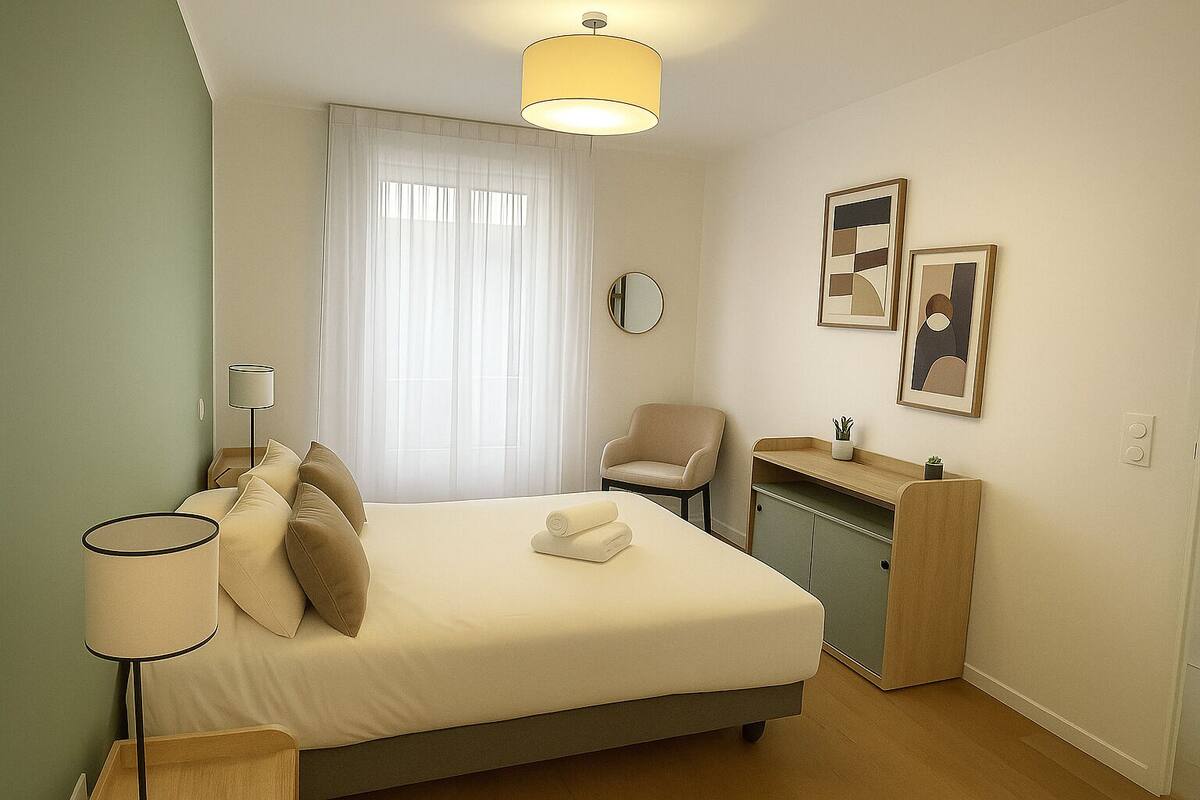 Een keurig opgemaakt slaapkamer met een bed, nachtlampjes, een stoel, een klein bureau met een plant, en twee ingelijste kunstwerken aan de muur.