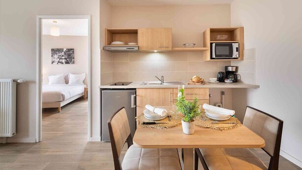 Een compacte hotelkamer met kitchenette, een eethoek met een tafel gedekt voor twee personen, en een bed met wit beddengoed.