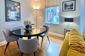 Una sala de estar moderna con una mesa redonda de vidrio, dos sillas grises, un sofá amarillo y una ventana con vistas a edificios.