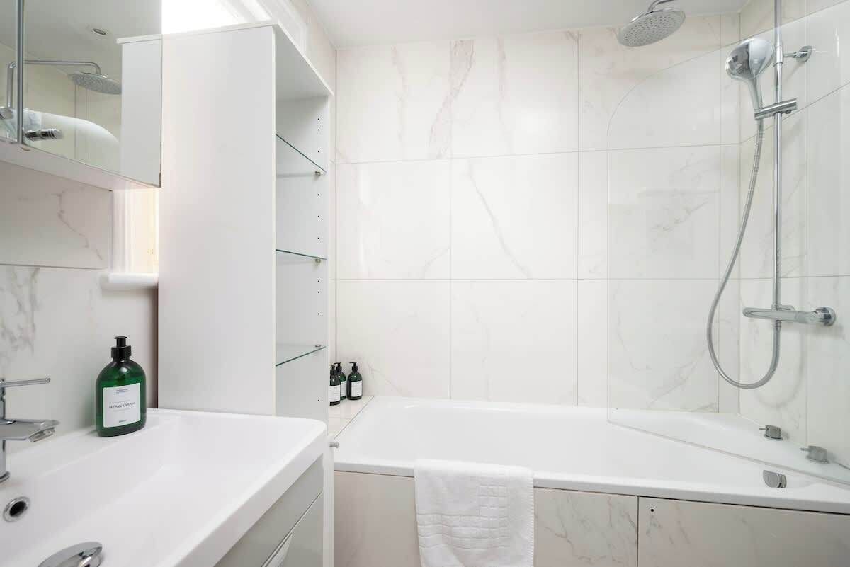 Een moderne badkamer met een witte marmeren wand, een badkuip, een douche met handdouche en een glazen plank met toiletartikelen.