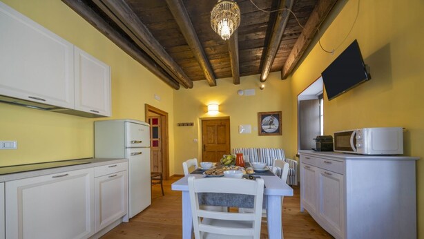 A kitchen with white cabinets, a dining table with chairs, a chandelier, and a television mounted on the wall. - GuestHost - A welcoming apartment of approximately 50 square meters, perfect for five people, located on the first floor of a historic building built in 1864 and renovated in 2025 while maintaining its historic atmosphere (NO elevator).The property is lo (Tarvisio)