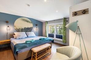 A bedroom with a bed, bench, chair, wall art, and a window with curtains.