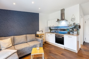 Een gezellig, compacte leefruimte met een bank, een klein tafeltje, een kitchenette met een kookplaat, vaatwasser en een klok aan de muur.