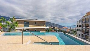 Area piscina moderna con un pontile in legno, ringhiera in vetro e vista sugli edifici e sulle montagne circostanti.
