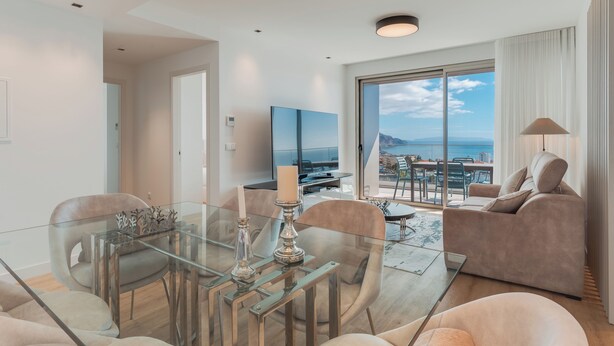 Un soggiorno moderno con un tavolo da pranzo in vetro, divani beige e un'ampia finestra con vista sul mare.