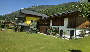 Un bâtiment à deux étages doté d’un balcon, entouré d’une pelouse bien entretenue et d’une colline boisée.