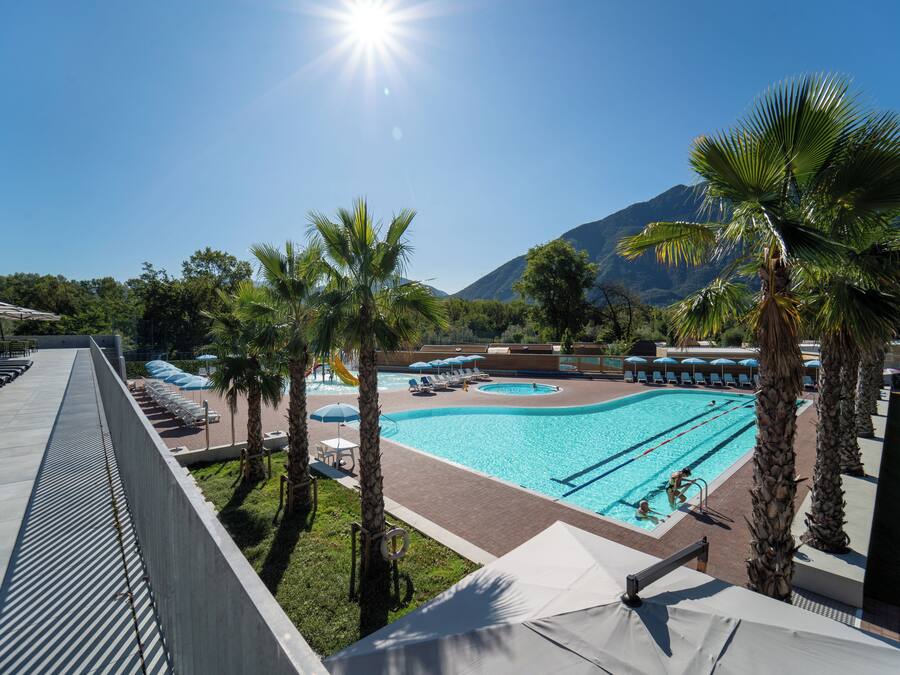 A swimming pool with lounge chairs and umbrellas, surrounded by palm trees and mountains in the background.