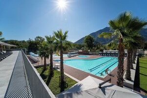 Piscina com espreguiçadeiras e guarda-sóis, cercada por palmeiras e montanhas ao fundo.