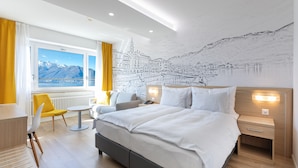 Ein modernes Hotelzimmer mit einem großen Bett, einem Schreibtisch, einem Sessel und Blick auf Berge durch das Fenster.