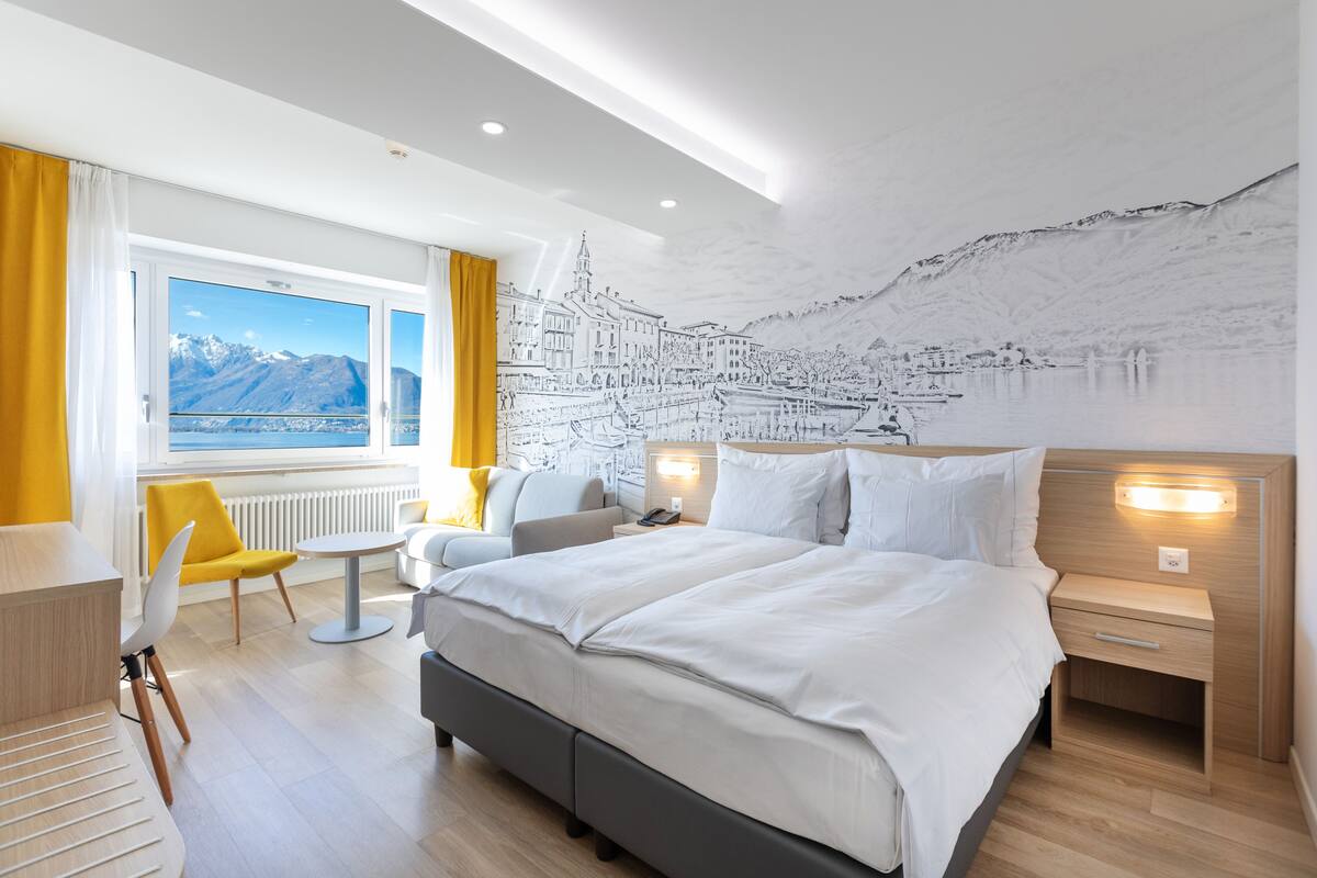 Une chambre d’hôtel moderne, dotée d’un grand lit, d’un bureau, d’une chaise et offrant une vue sur les montagnes par la fenêtre.