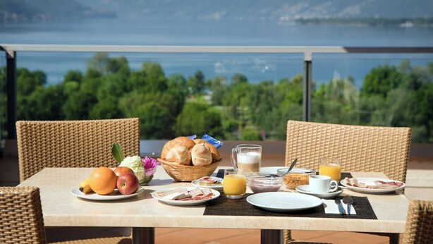Ein Esstisch mit Speisen, einem Brotkorb, einer Saftkanne und einer Kaffeetasse, platziert auf einem Balkon mit Blick auf einen See und Berge.