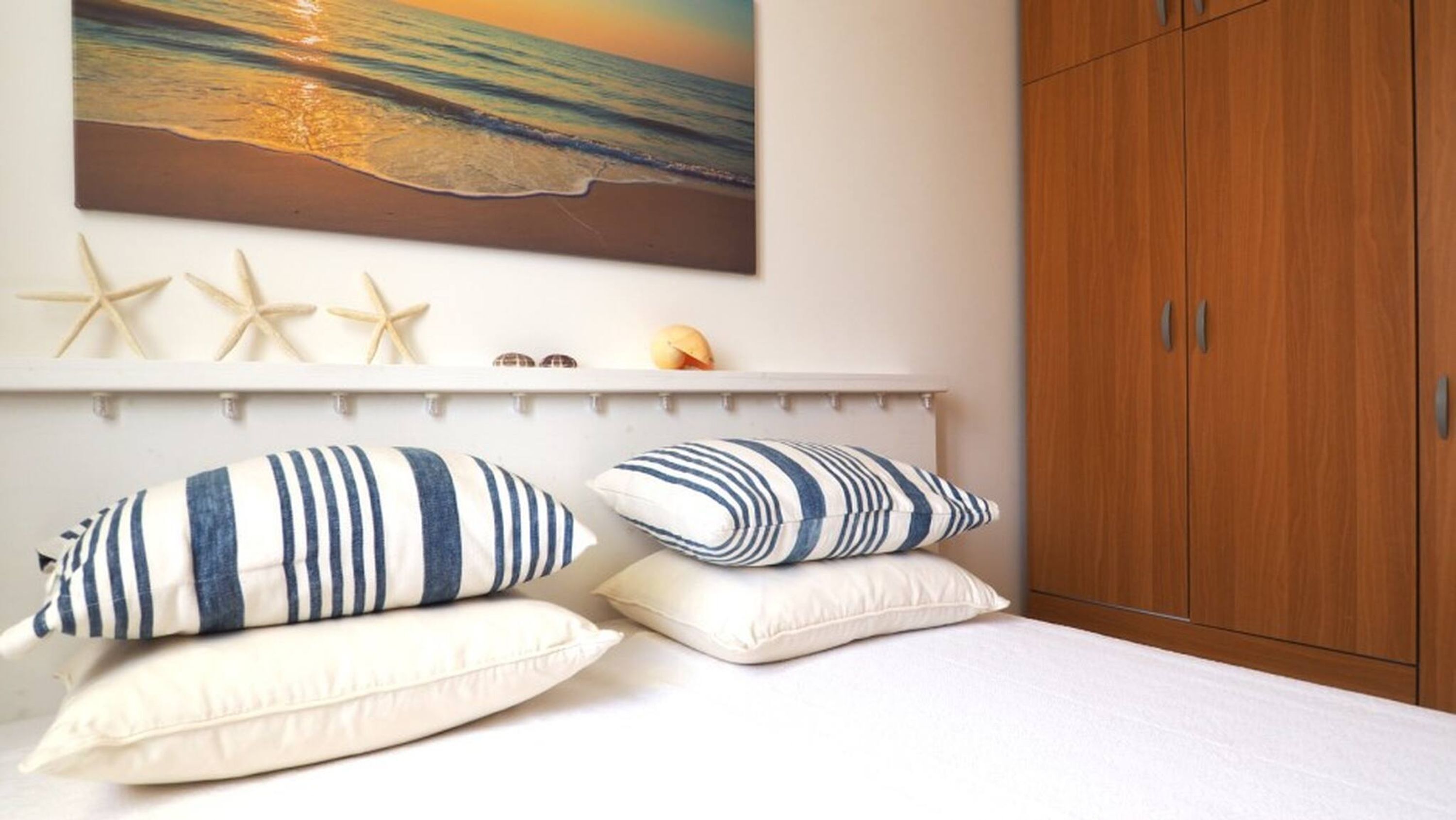 Ein Schlafzimmer mit einem Bett, gestreifte Kissen, einem an der Wand befestigten Regal mit Seesternen und Muscheln sowie einem Holzschrank.