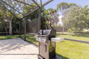 An outdoor grill area with a metal grill, a sliding glass door, and a view of a grassy area with trees. - Stylish 3BR w/ Heated Pool · 7 Mins to Beach (Jupiter)