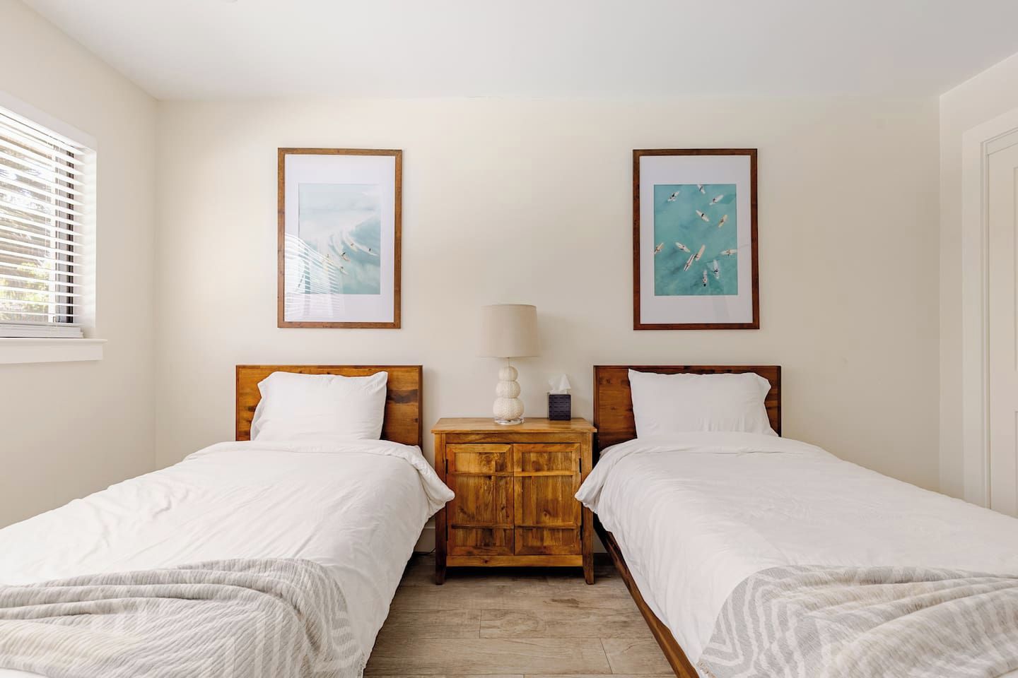 A hotel room with two single beds, a wooden nightstand with a lamp, and two framed pictures on the wall.