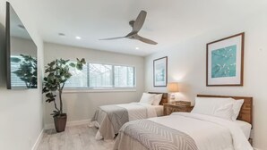 Quarto com duas camas, ventilador de teto, planta em vaso e obras de arte nas paredes.
