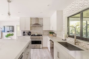 Une cuisine moderne avec des meubles blancs, un four encastré, une cuisinière en acier inoxydable et une grande fenêtre donnant sur un paysage verdoyant.