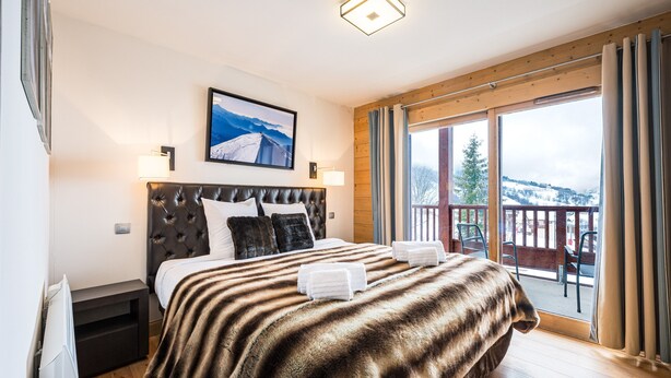 A bedroom with a large bed, a headboard, a mounted picture, a nightstand, and a view of the outdoors.