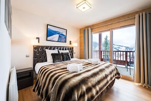 A bedroom with a large bed, a headboard, a mounted picture, a nightstand, and a view of the outdoors.