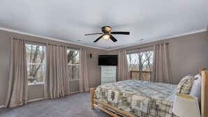 Un dormitorio con cama, ventilador de techo, televisor y ventanas grandes con cortinas.