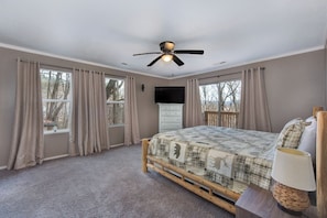 Ein Schlafzimmer mit einem Bett, einem Deckenventilator, einem Fernseher und großen Fenstern mit Vorhängen.