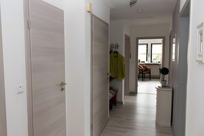 Un couloir avec une porte en bois de couleur claire, un porte-manteau sur lequel est accroché un blouson vert, une chaise et une petite table sur laquelle se trouve un objet décoratif.