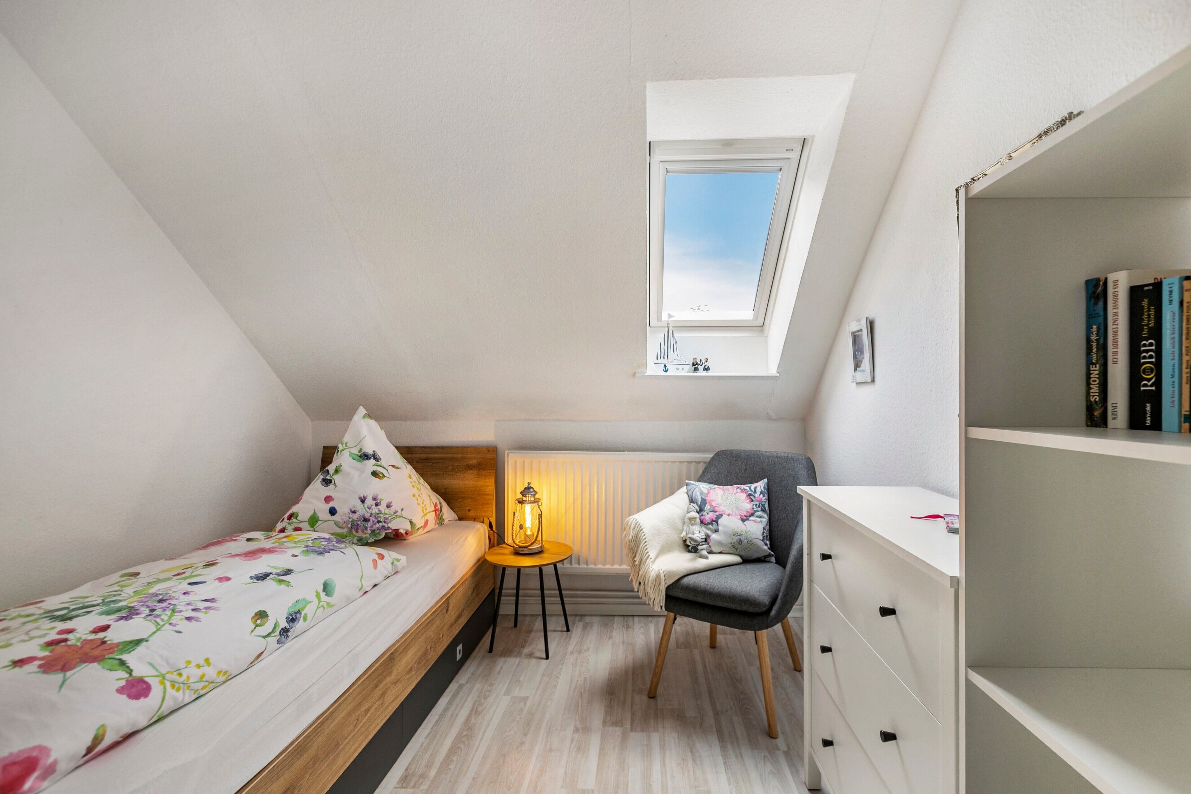 Una camera compatta con un letto singolo, un tavolino con una lampada, una sedia e una libreria.