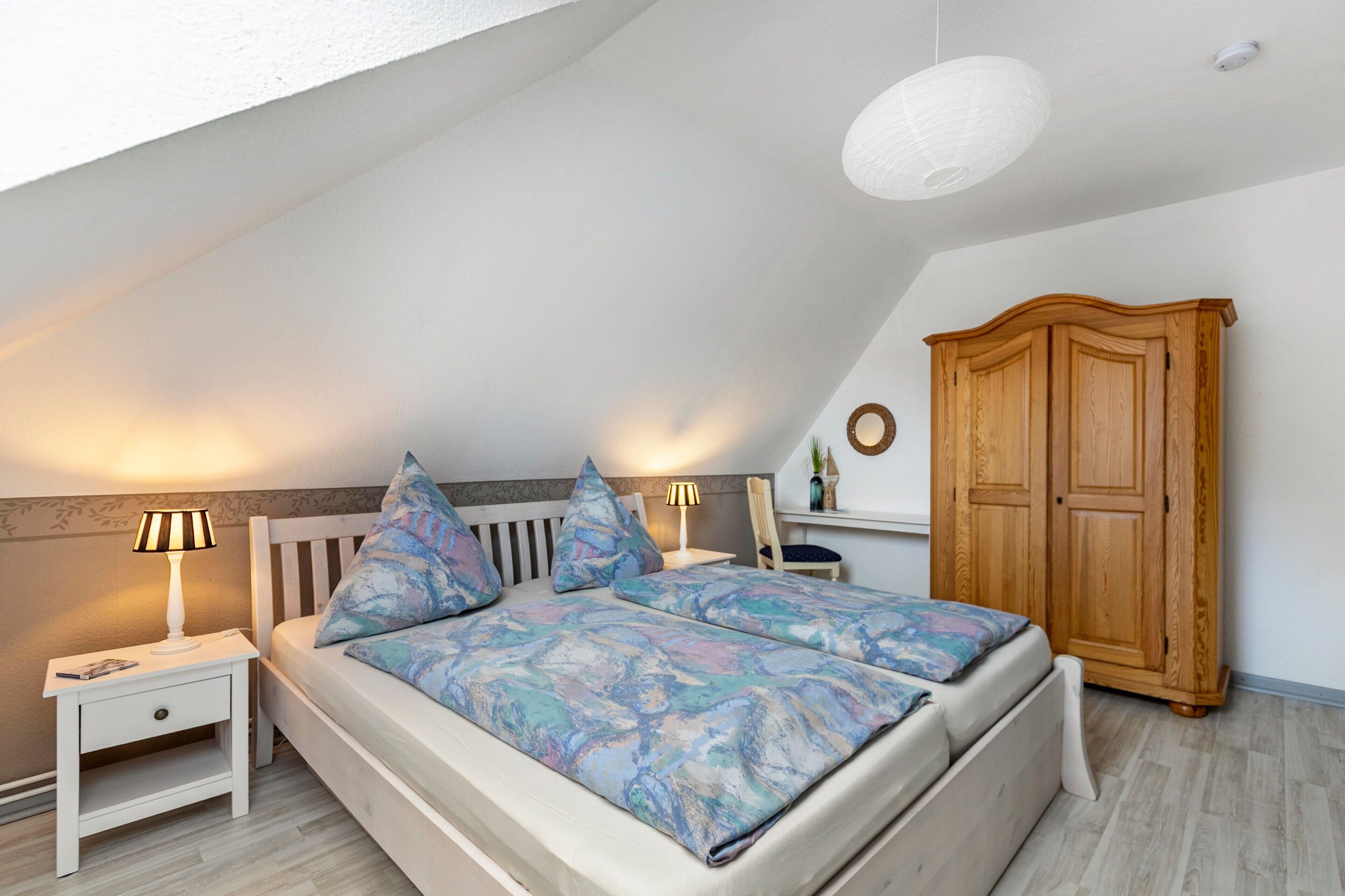Une chambre avec un plafond en pente, un lit recouvert d’une couvre-lit à motifs, des lampes de chevet, une armoire en bois et une petite table avec une lampe.