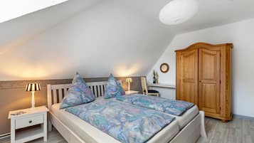 Une chambre avec un plafond en pente, un lit recouvert d’une couvre-lit à motifs, des lampes de chevet, une armoire en bois et une petite table avec une lampe.