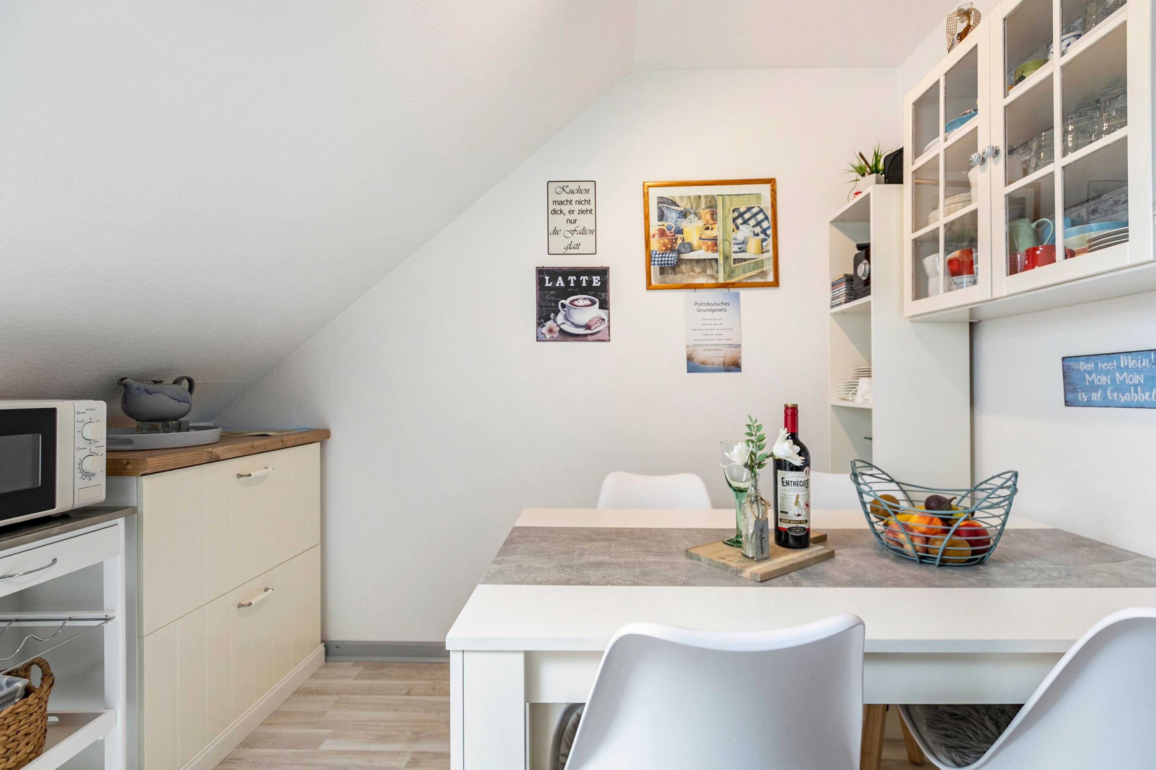 Et moderne køkken med hvide skabe, en mikrobølgeovn, et spisebord med stole og et glaskabinet med service.