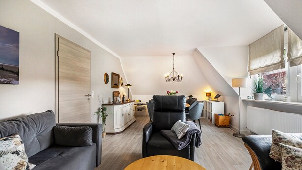 Ein modernes Wohnzimmer mit Sofa, Sessel und Couchtisch. Im Hintergrund ist eine Küchenzeile mit Esstisch und Stühlen zu sehen.