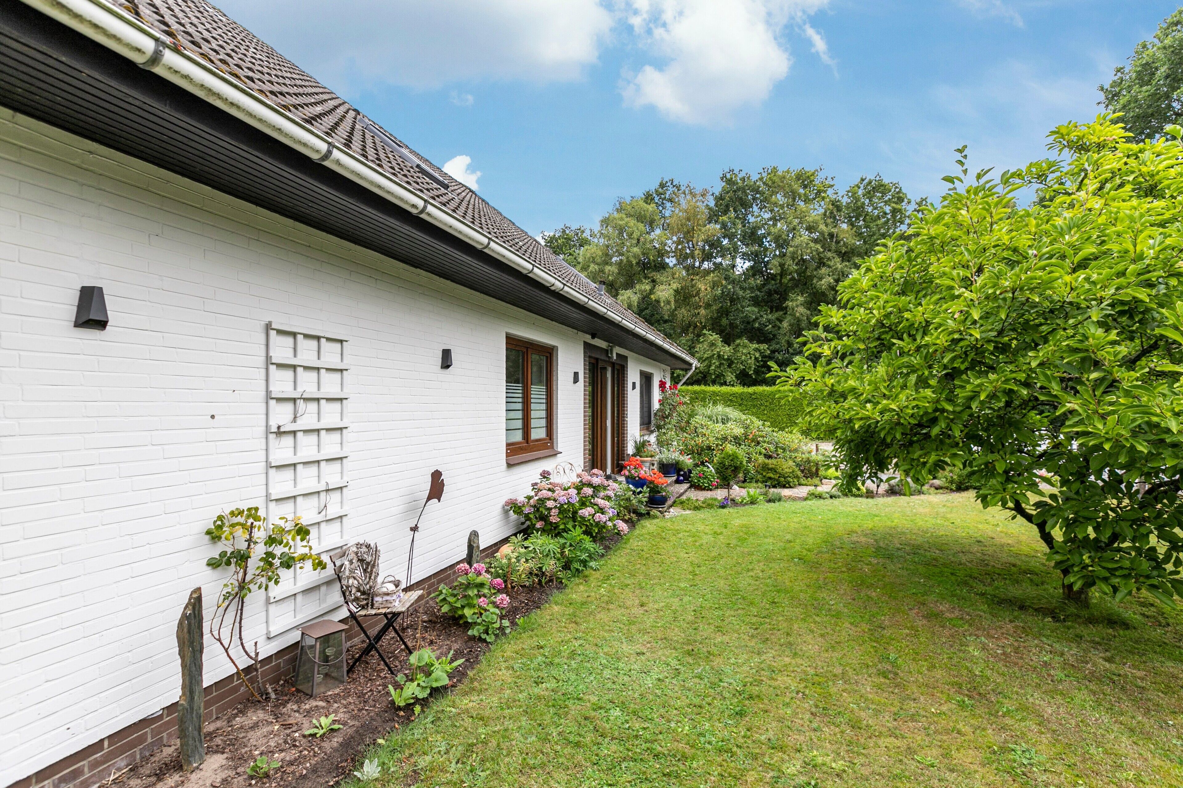Ein weißes Haus mit geneigtem Dach, einem Backsteinfundament und einem gepflegten Garten mit verschiedenen Pflanzen und Blumen.