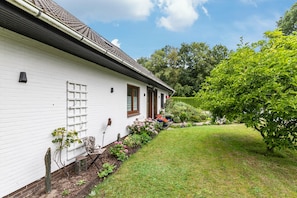 Et hvidt hus med skråt tag, murstenssokkel og en velplejet have med forskellige planter og blomster.