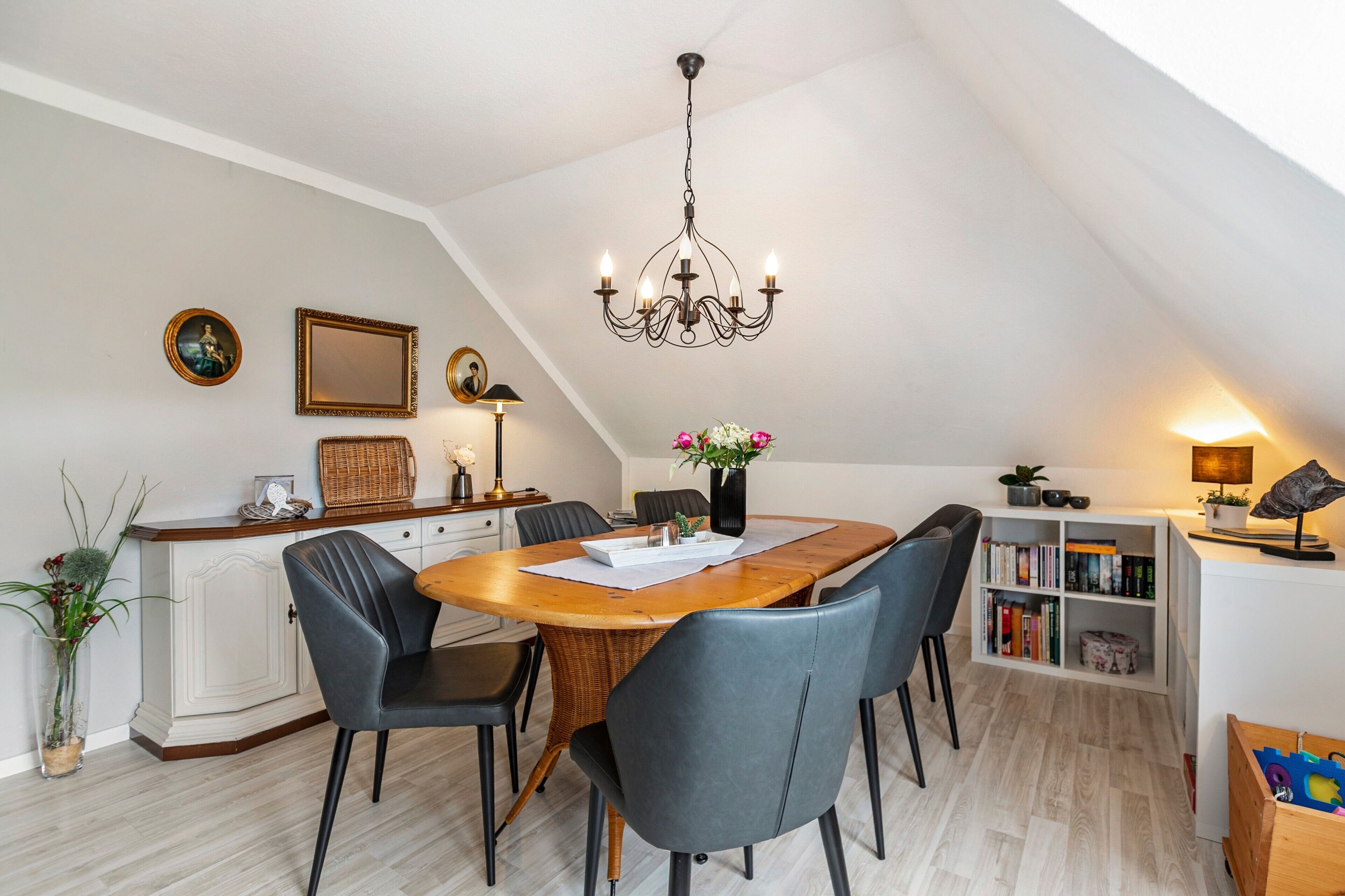 Un coin repas avec une table en bois, des chaises à revêtement gris, un lustre et une bibliothèque.
