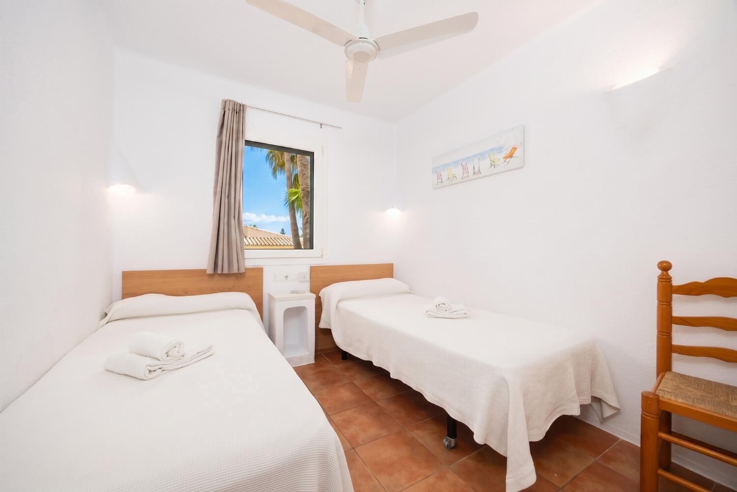 A hotel room with two beds, a ceiling fan, a window with a view, a small table, and a chair.