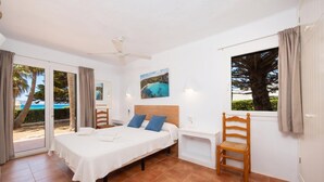 A bedroom with a bed, a ceiling fan, a window with curtains, and a view of the beach.