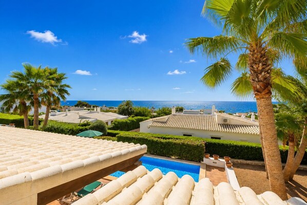 A pool area with a clear view of the sea, surrounded by palm trees and a tiled roof.