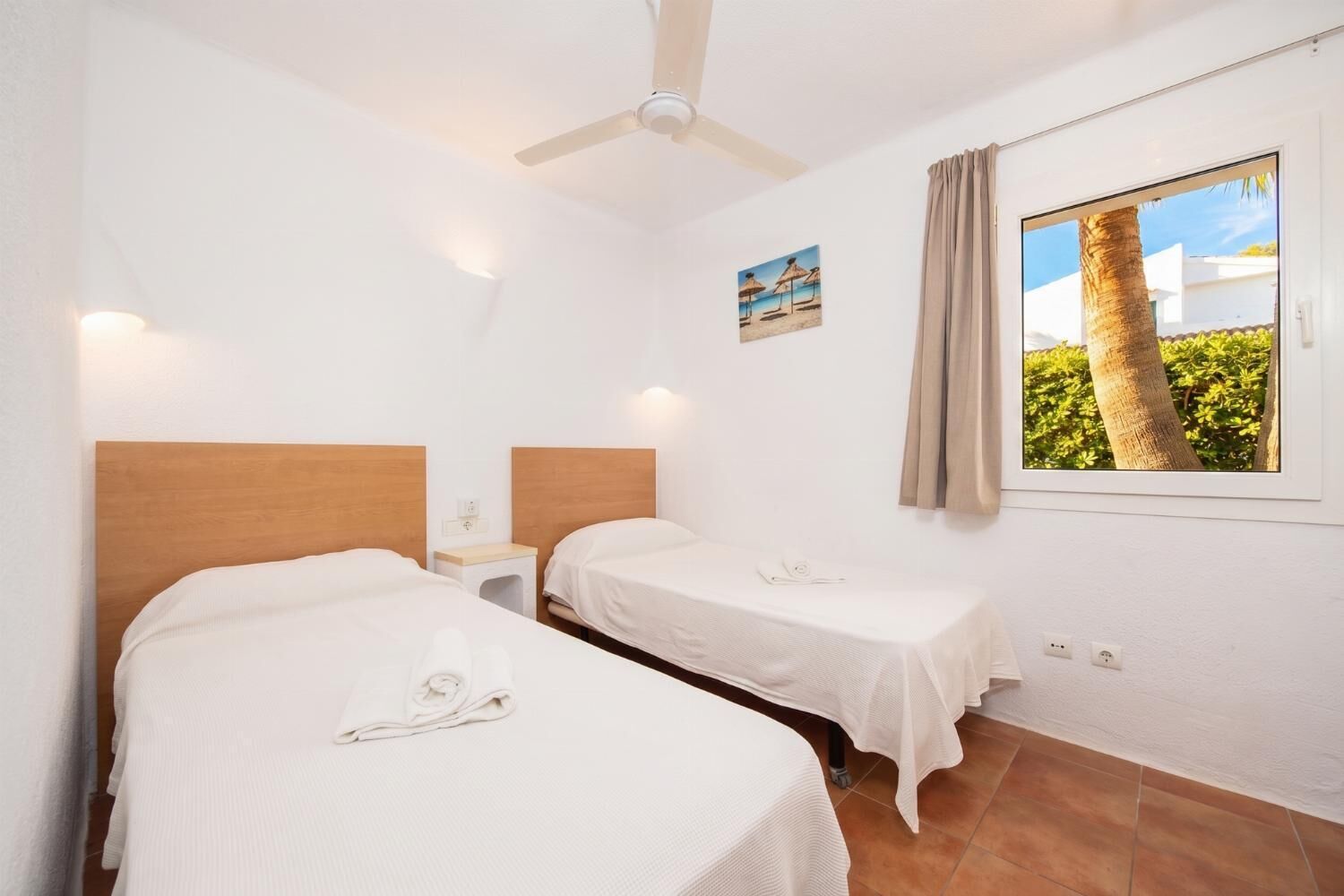 Una stanza con due letti, un ventilatore a soffitto, una finestra vista verde e un quadro appeso al muro.