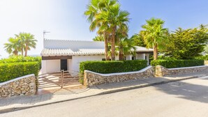 A white villa with a tiled roof, surrounded by palm trees and hedges, featuring a stone pathway leading to the entrance.