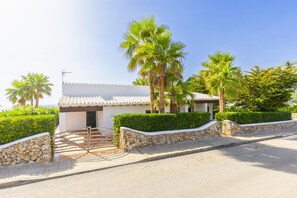 A white villa with a tiled roof, surrounded by palm trees and hedges, featuring a stone pathway leading to the entrance.