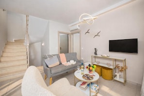 A modern living room with a staircase, a grey sofa, a coffee table with books and a fruit bowl, a TV, and a minimalist decor. - Villa Aurora - Three Bedroom Villa, Sleeps 6 (Mparmpati)