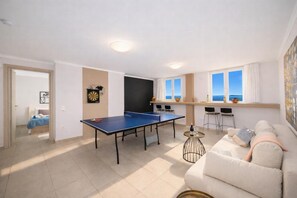 A room with a ping pong table, a sofa, a dining area with chairs, and a view of the sea through large windows. - Villa Aurora - Three Bedroom Villa, Sleeps 6 (Mparmpati)