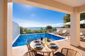 Un espace repas aménagé au bord de la piscine, avec une table dressée pour deux personnes, une bouteille de vin et un verre d’eau.