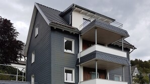 A multi-story residential building with a dark exterior, white trim, and a balcony on the upper floor. - Apartment "Olsberg View Whg. B" with Mountain View, Shared Garden and Wi-Fi (Olsberg)