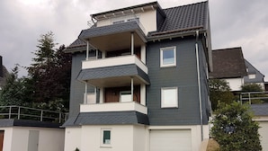 A multi-story residential building with a flat roof, a balcony on the second floor, and a garage door. - Apartment "Olsberg Blick Whg.A" with Mountain View, Garden & Wi-Fi (Olsberg)