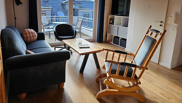 A modern living room with a sofa, a wooden rocking chair, a dining table, and a balcony with a view of buildings and mountains. - Apartment "Olsberg Blick Whg.A" with Mountain View, Garden & Wi-Fi (Olsberg)