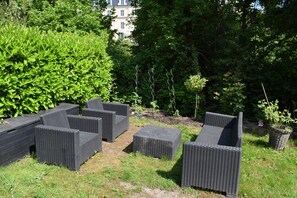 Gartenmöbel aus Rattan im Freien, umgeben von üppigem Grün und einem Gebäude im Hintergrund.